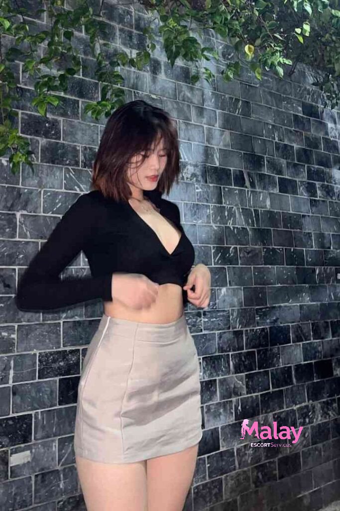 Malay Escort Service Young woman in a black crop top and satin skirt stands against a brick wall KL Escort Girl