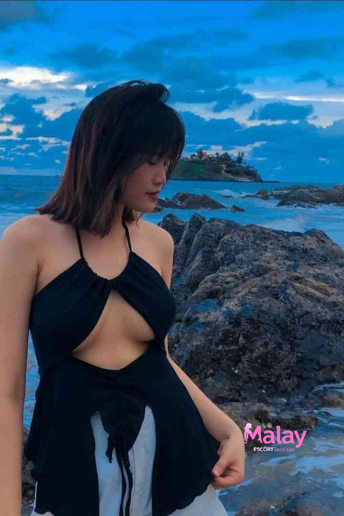 Malay Escort Service Woman in black halter top on rocky beach with cloudy blue sky KL Escort Girl