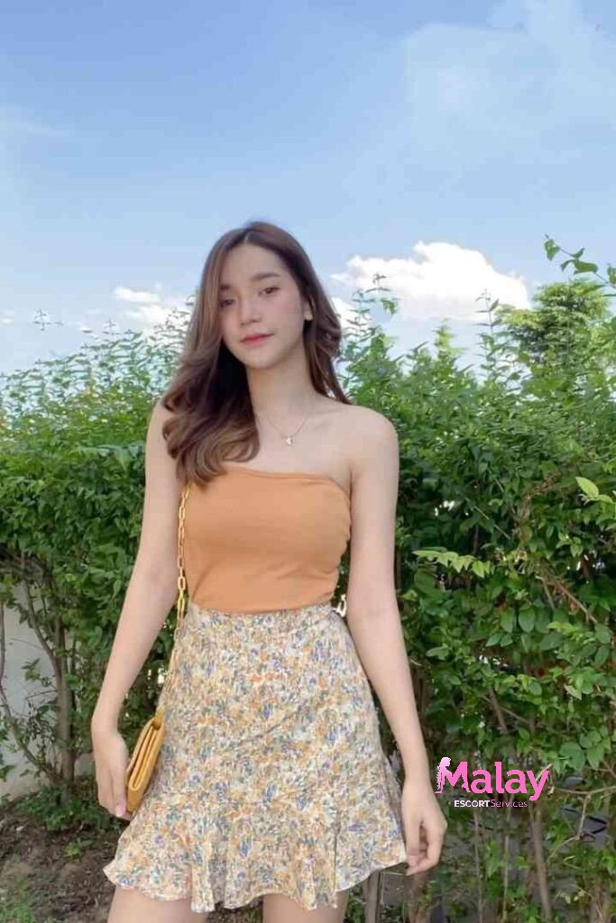 Malay Escort Service Young woman in a floral skirt and orange tube top poses outdoors with a green hedge background KL Escort Girl