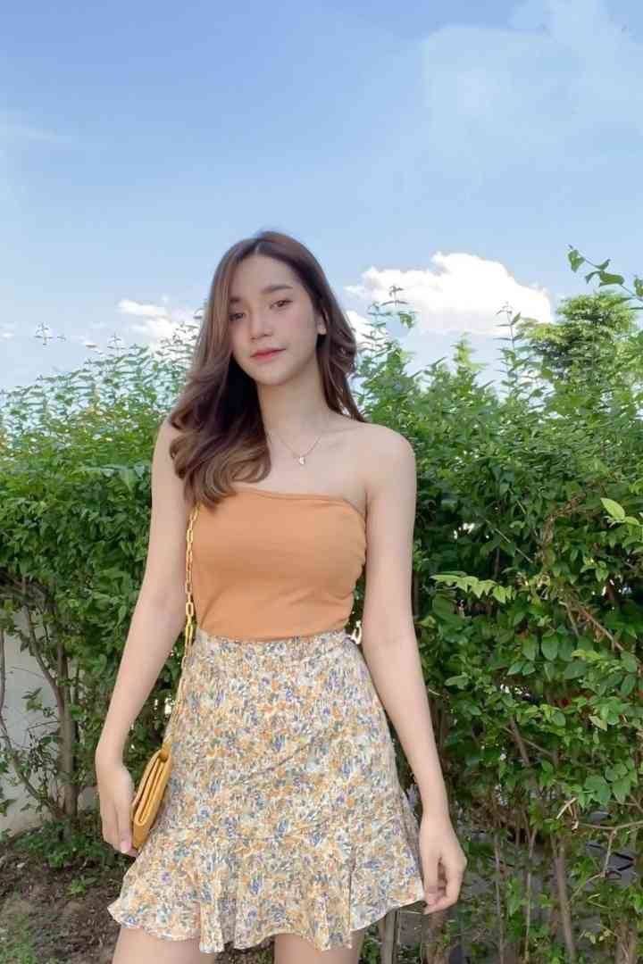 Malay Escort Service Young woman in a floral skirt and orange tube top standing outdoors with lush green foliage. KL Escort Girl