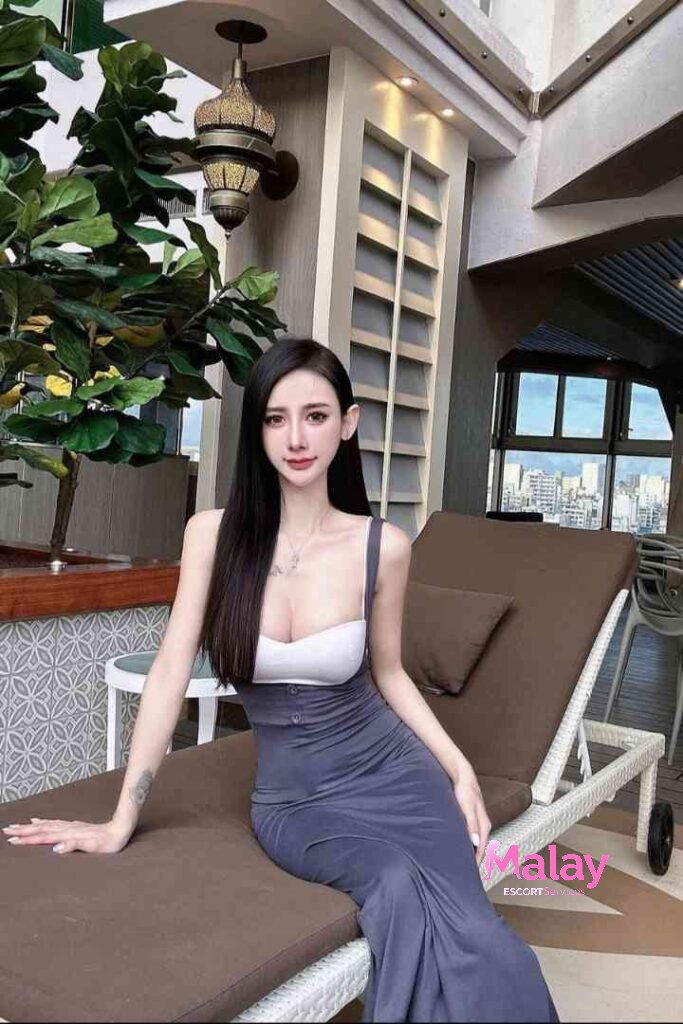 Malay Escort Service Woman in a gray dress relaxing on a lounge chair with lush green plants and city view KL Escort Girl Malay Escort KL Real KL Escorts Ready for You Malay Escort Service Woman in a gray dress relaxing on a lounge chair with lush green plants and city view KL Escort Girl