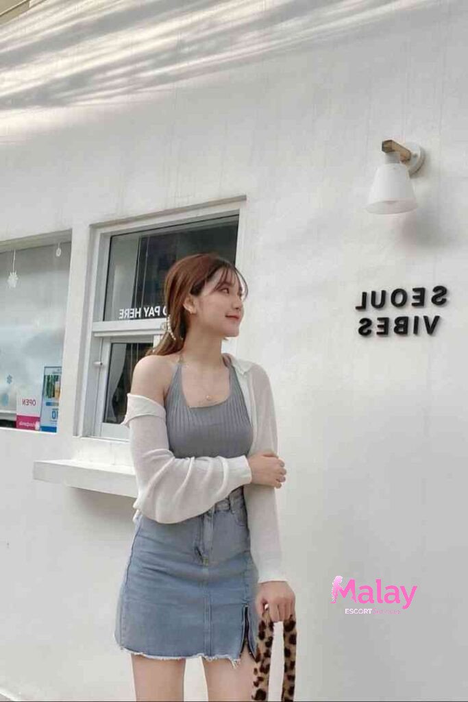 Malay Escort Service Young woman in a grey halter top and denim skirt stands against a white wall with text KL Escort Girl