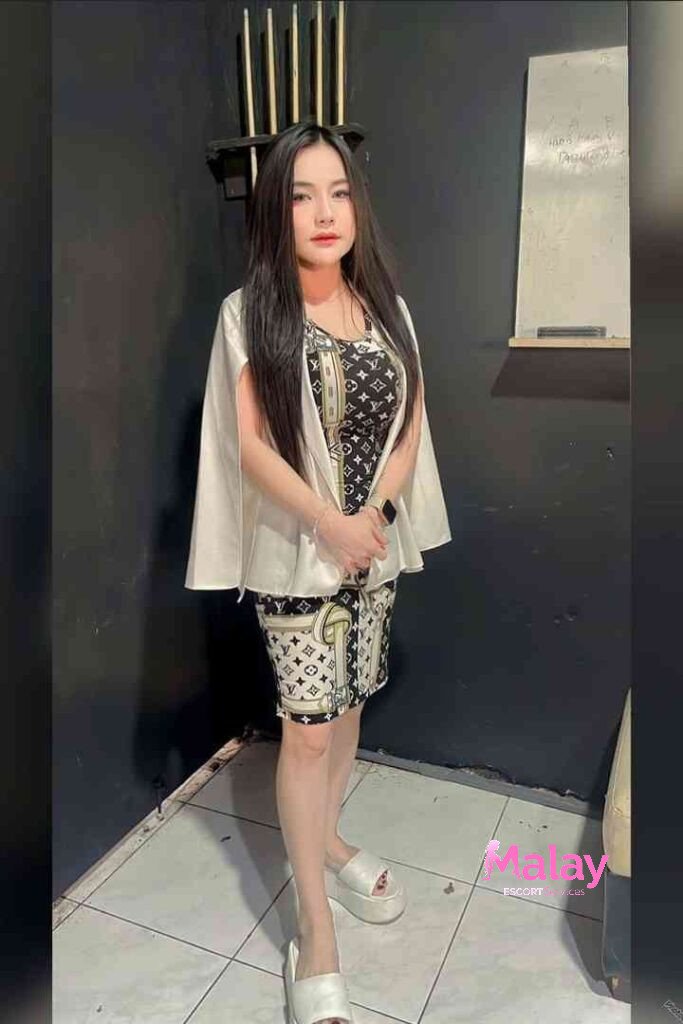 Malay Escort Service Woman in a patterned dress and white capelet stands against a dark wall KL Escort Girl