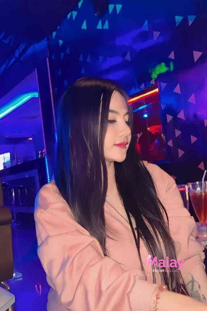 Malay Escort Service Young woman with long dark hair and highlights in a pink jacket at a bar with blue and purple lighting KL Escort Girl