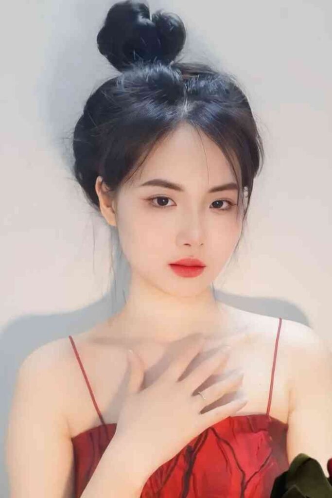 Malay Escort Service Young woman with dark hair in a bun wearing a red spaghetti strap dress with her hand on her chest KL Escort Girl