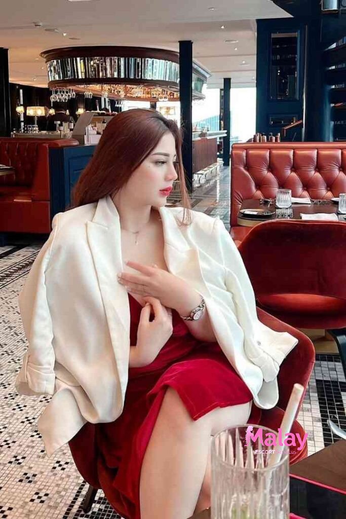 Malay Escort Service Woman in red dress and white blazer sits in a restaurant booth KL Escort Girl