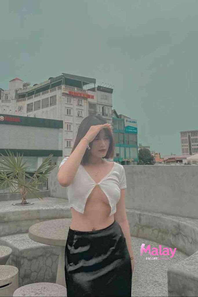 Malay Escort Service Young woman in a white crop top and black skirt stands on a rooftop with buildings in the background KL Escort Girl