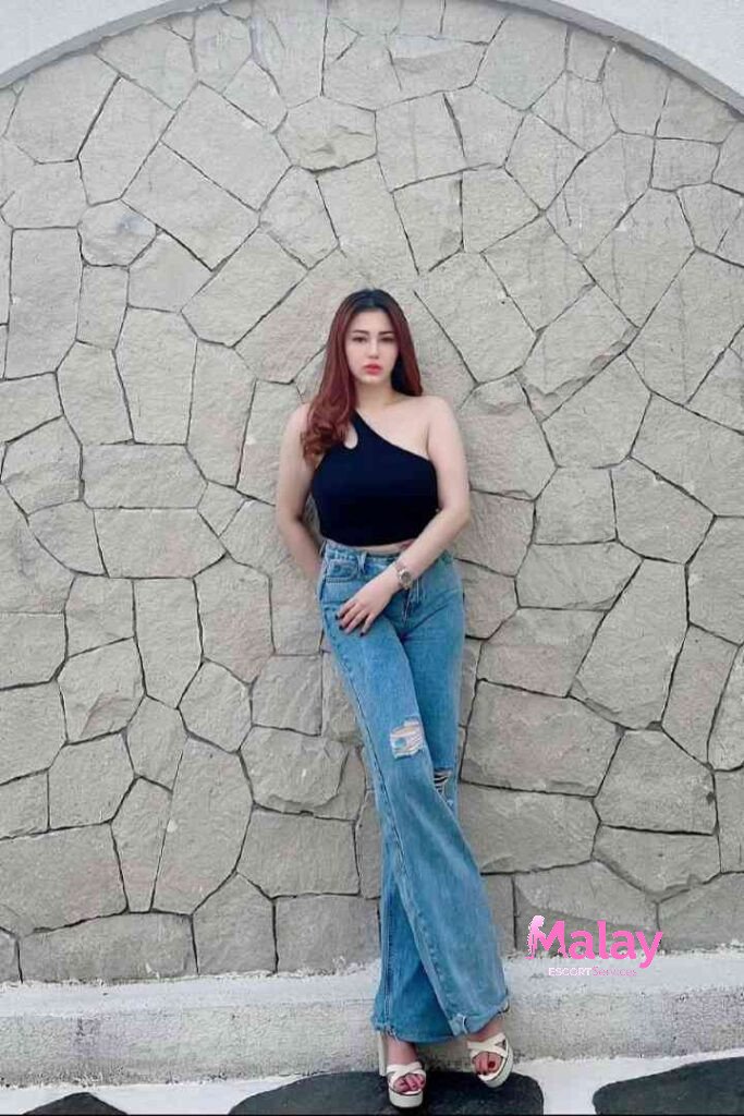 Malay Escort Service Woman in ripped jeans and a black one shoulder top leans against a stone wall KL Escort Girl