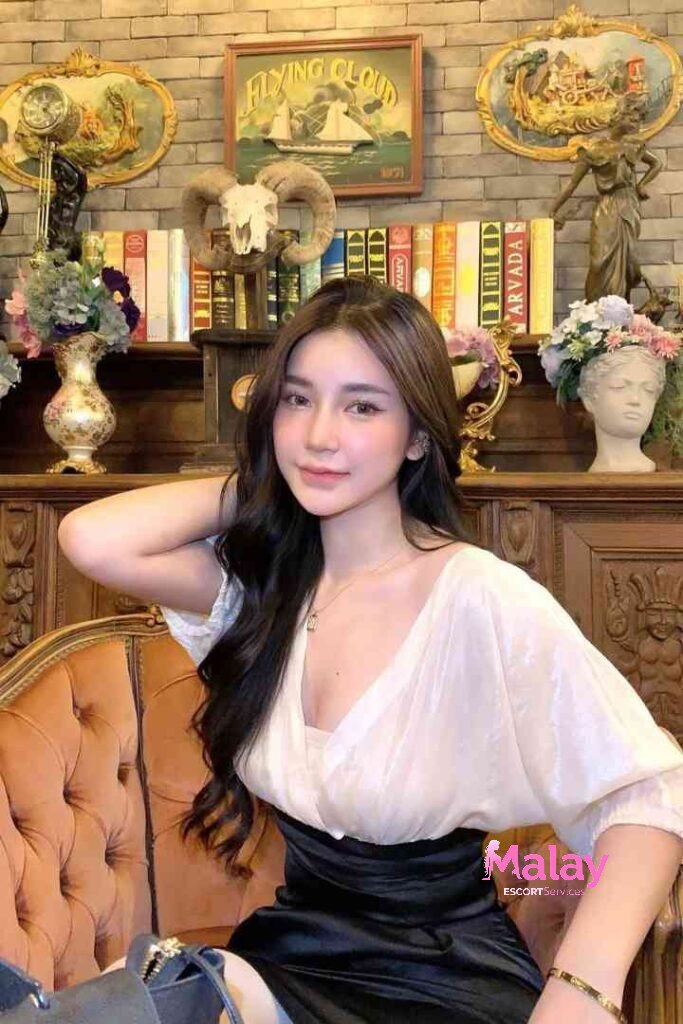 Malay Escort Service Young woman with long dark hair wearing a white top and black skirt sitting on an ornate couch KL Escort Girl