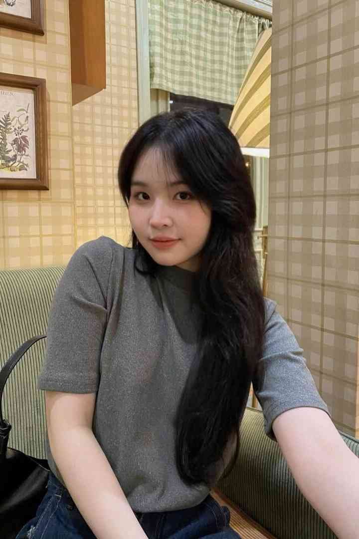 Malay Escort Service Young woman with long black hair wearing a grey t-shirt sits on a couch. KL Escort Girl