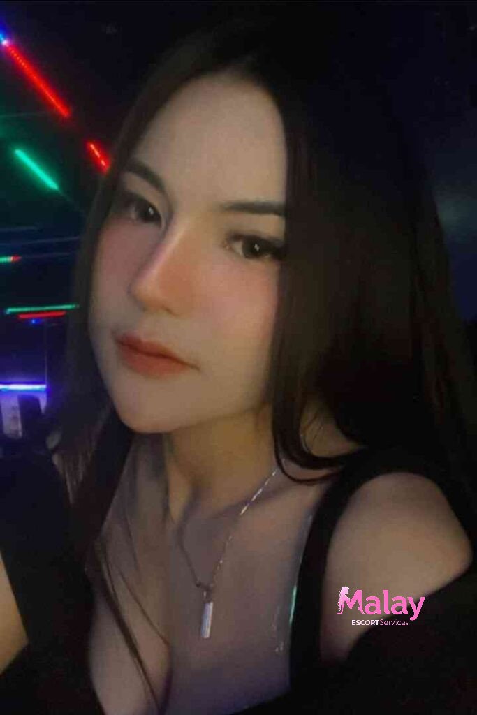 Malay Escort Service Young woman with long dark hair and a necklace softly lit with colorful lights in the background KL Escort Girl