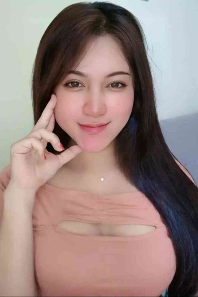 Young woman with long dark hair and light brown eyes wearing a pink top poses with her hand on her chin