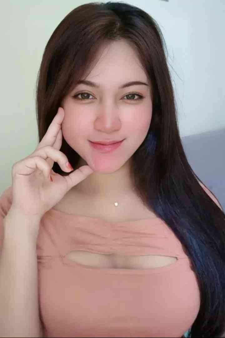 Young woman with long dark hair and light brown eyes, wearing a pink top, poses with her hand on her chin.