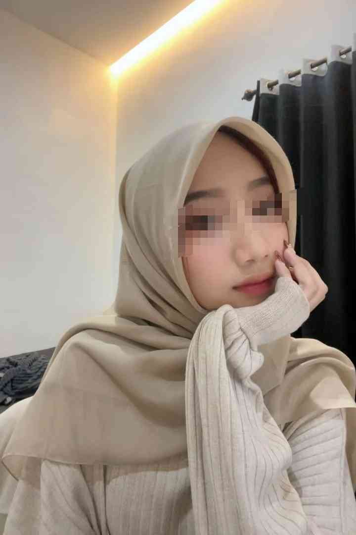 Malay Escort Service Young woman in a beige hijab and sweater, resting her chin on her hand. KL Escort Girl