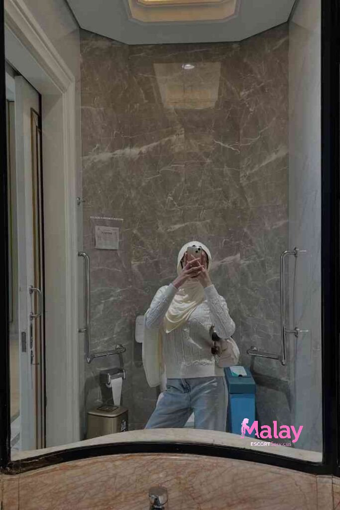 Malay Escort Service Woman in hijab and white sweater taking mirror selfie in modern bathroom KL Escort Girl
